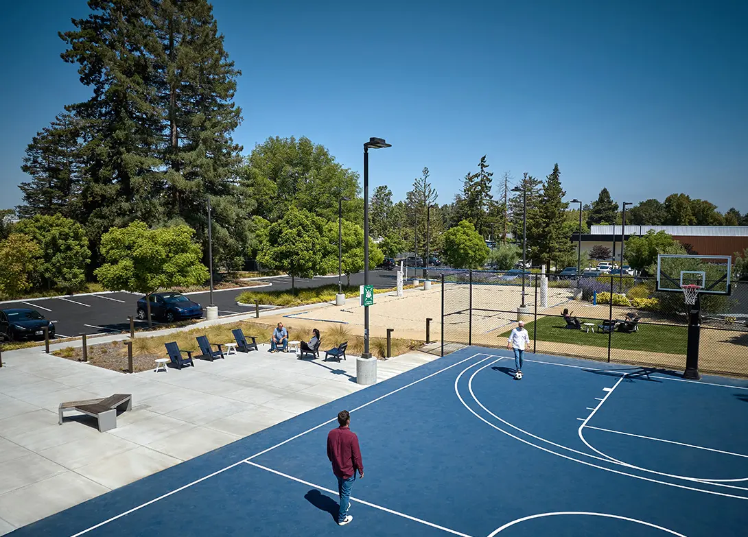 Outdoor basketball court with two players, surrounded by trees and seating area under clear skies.