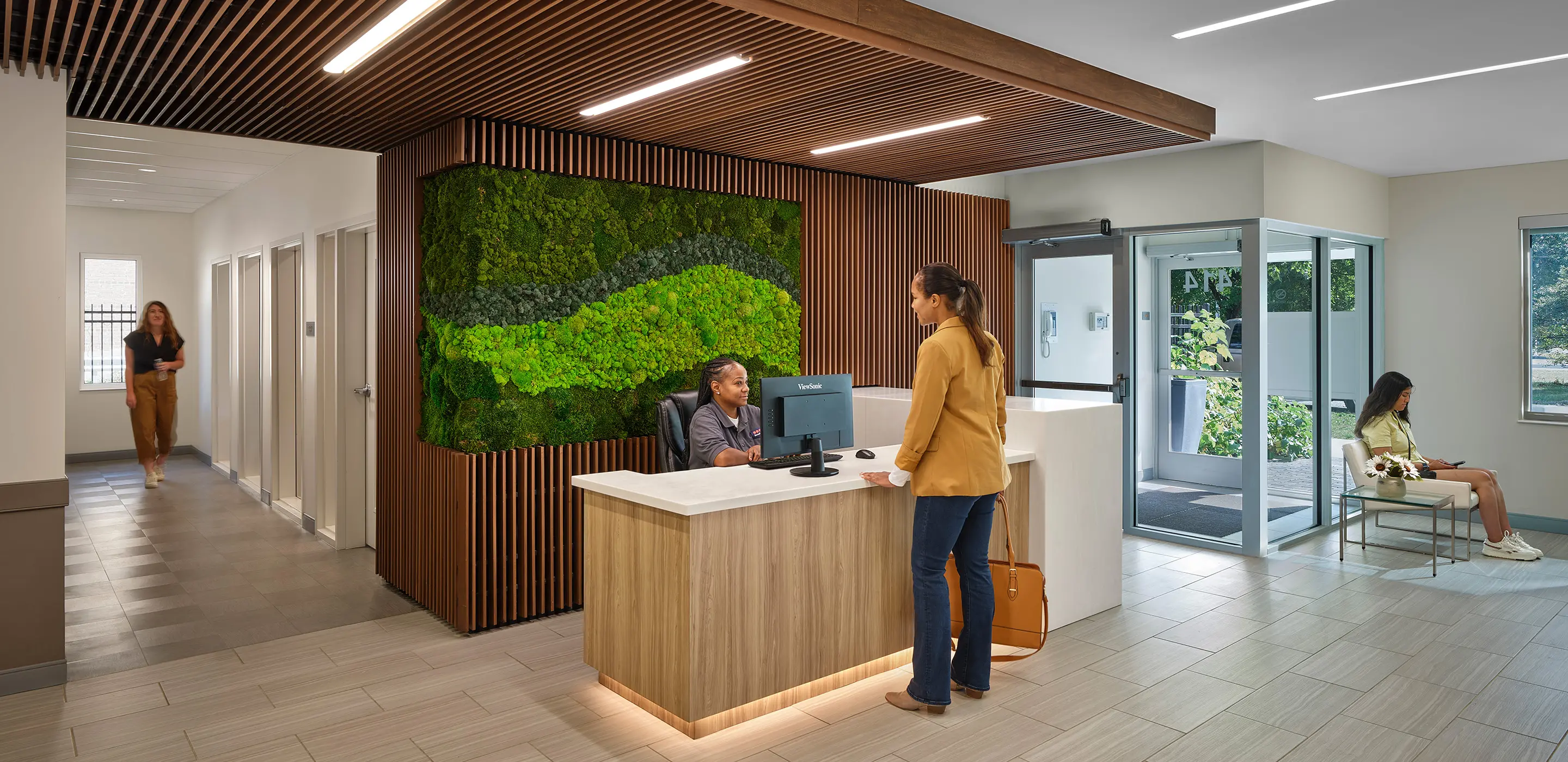 Modern reception area with green wall, receptionist at desk, and visitors in a waiting room with natural lighting.