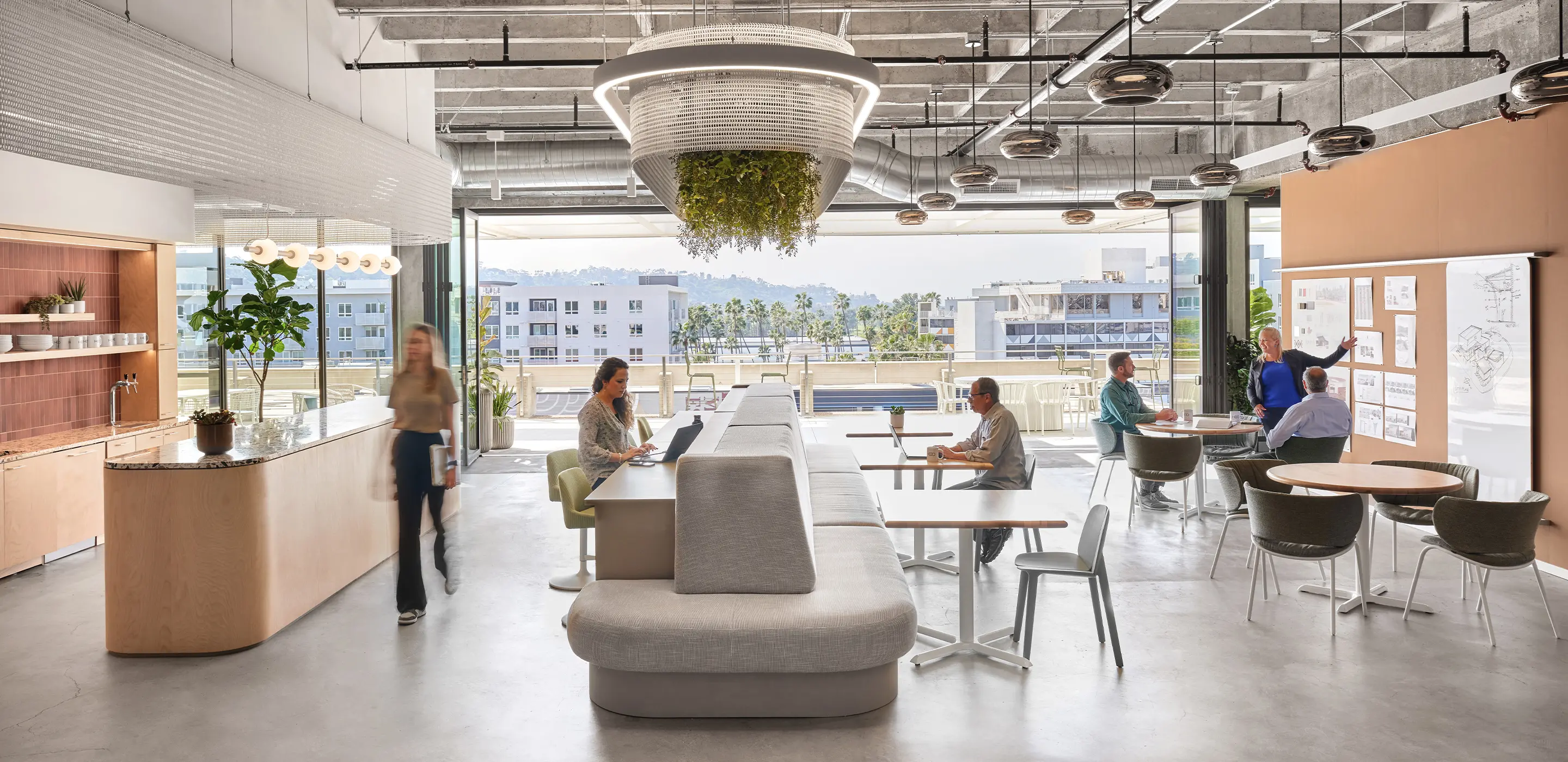 Modern coworking space with people collaborating, featuring open layout, greenery, and urban view through large windows.