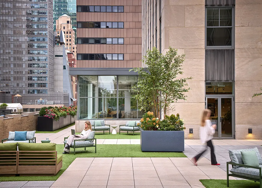 Rooftop garden with seating, plants, and city view. A woman on a laptop and another walking with a drink.