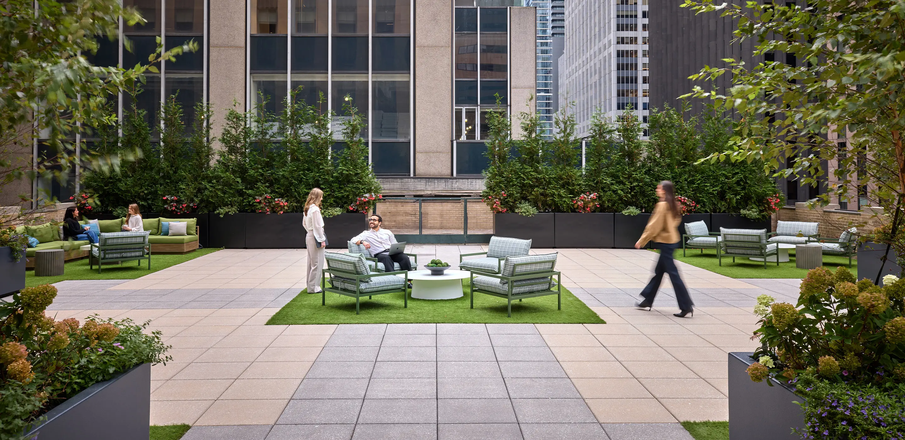 Outdoor office terrace with people interacting on cozy seating, surrounded by greenery and urban skyscrapers.