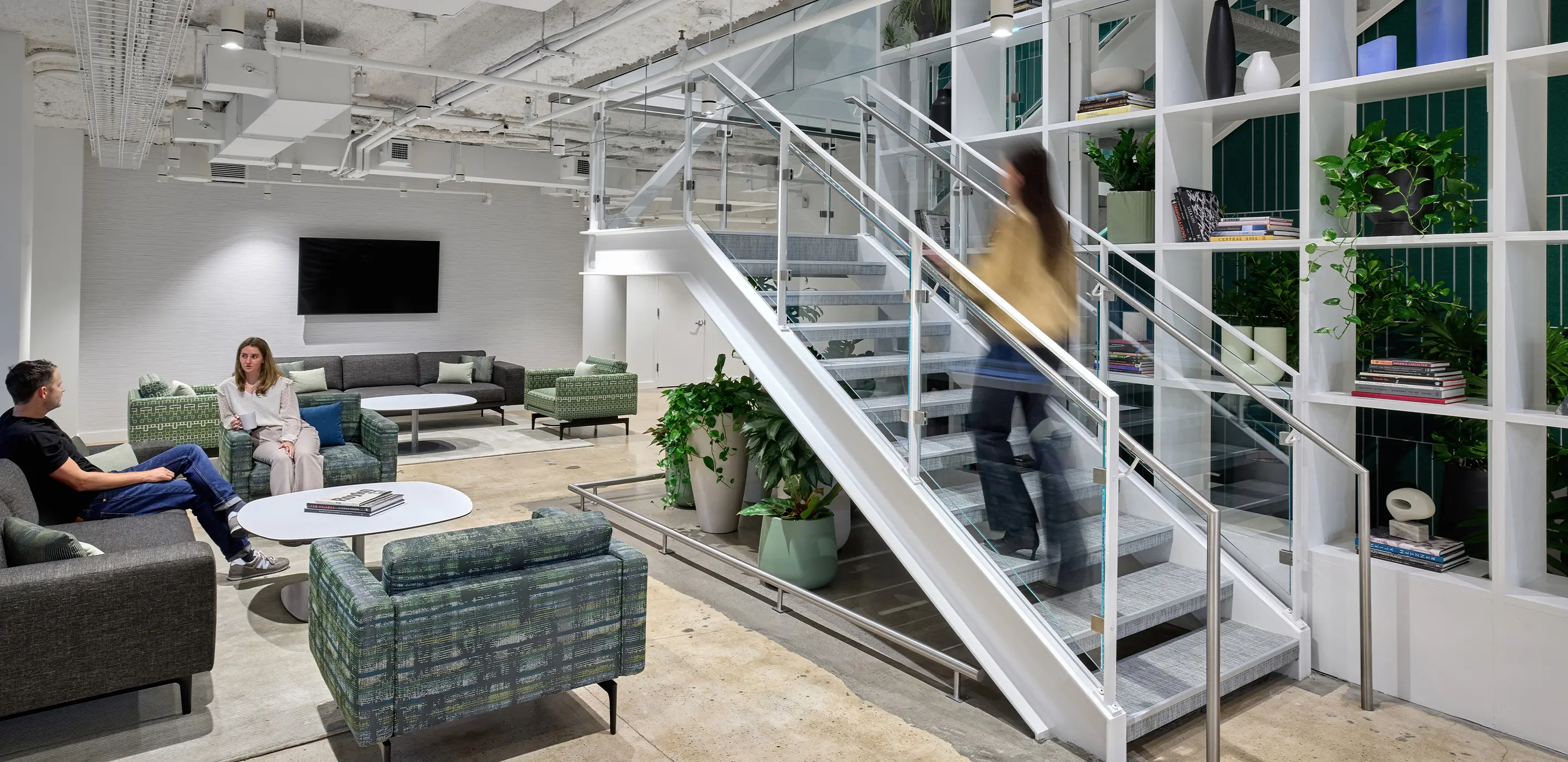 Modern office lounge with stairs, two people conversing on sofas, and greenery for a fresh, open atmosphere.