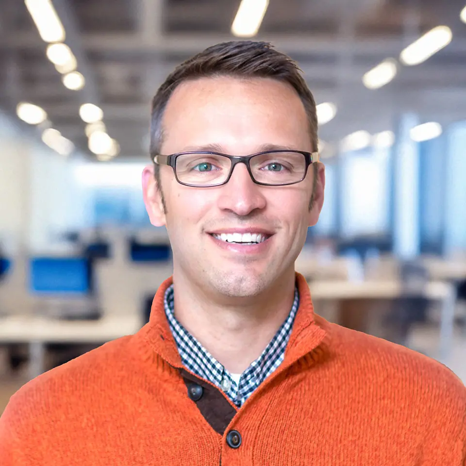 Smiling man in glasses and orange sweater in a modern office environment.
