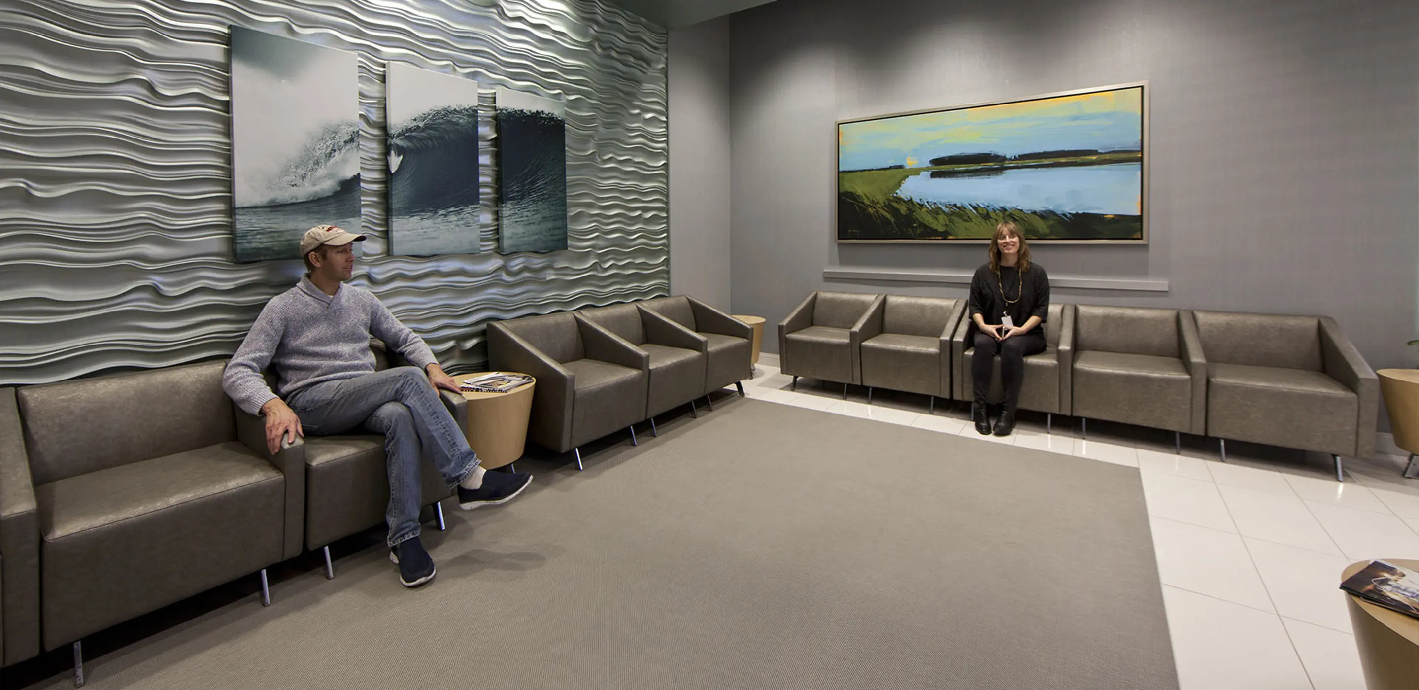 Modern waiting room with two people sitting on gray chairs, art on textured walls, and a calm, neutral design.