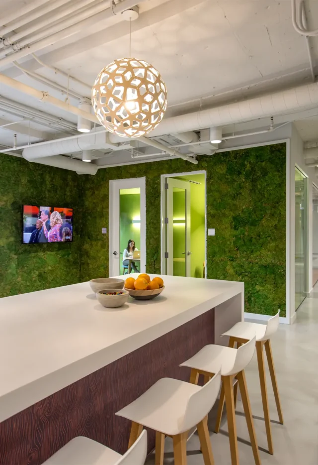 Modern office space with green walls, sleek pendant light, and white counter with fruit bowl and stools.