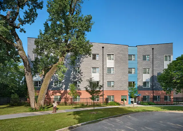 Four-story apartment building with modern facade and surrounding trees on a sunny day.