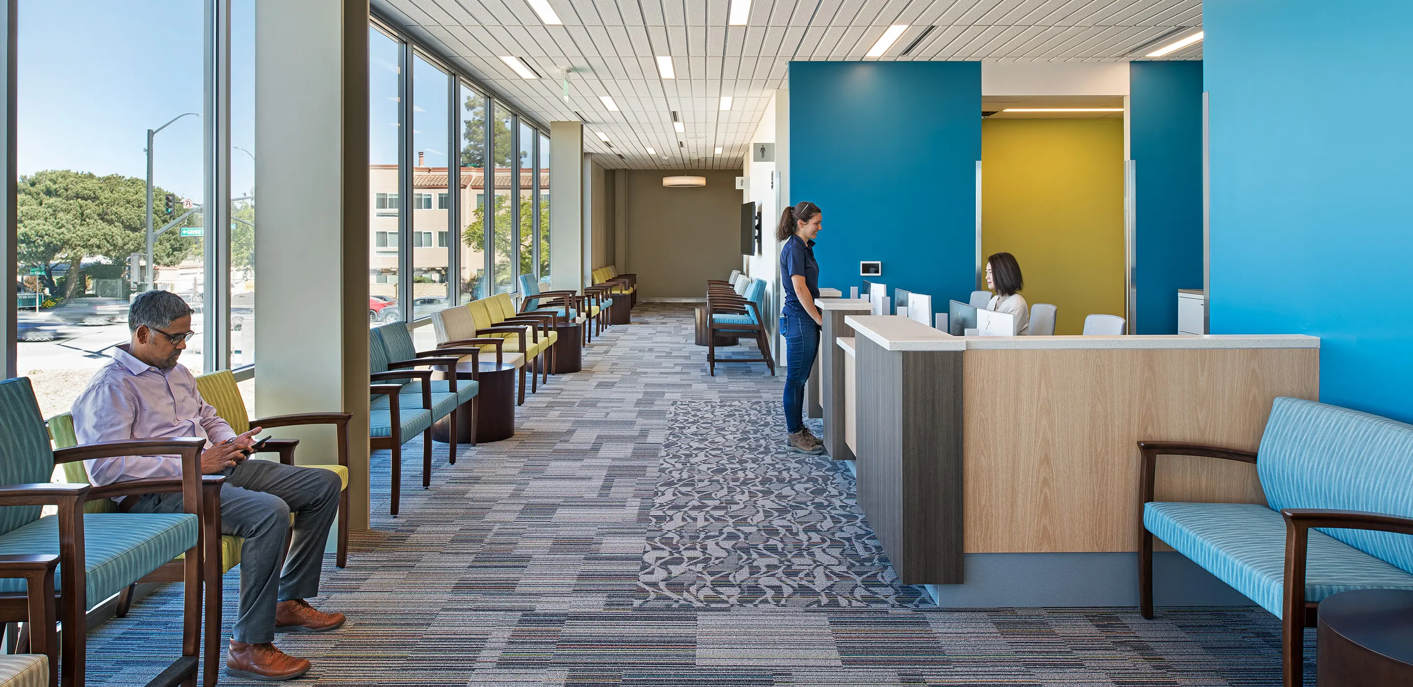 Modern waiting room with people seated and interacting at reception desk in a bright, open space.