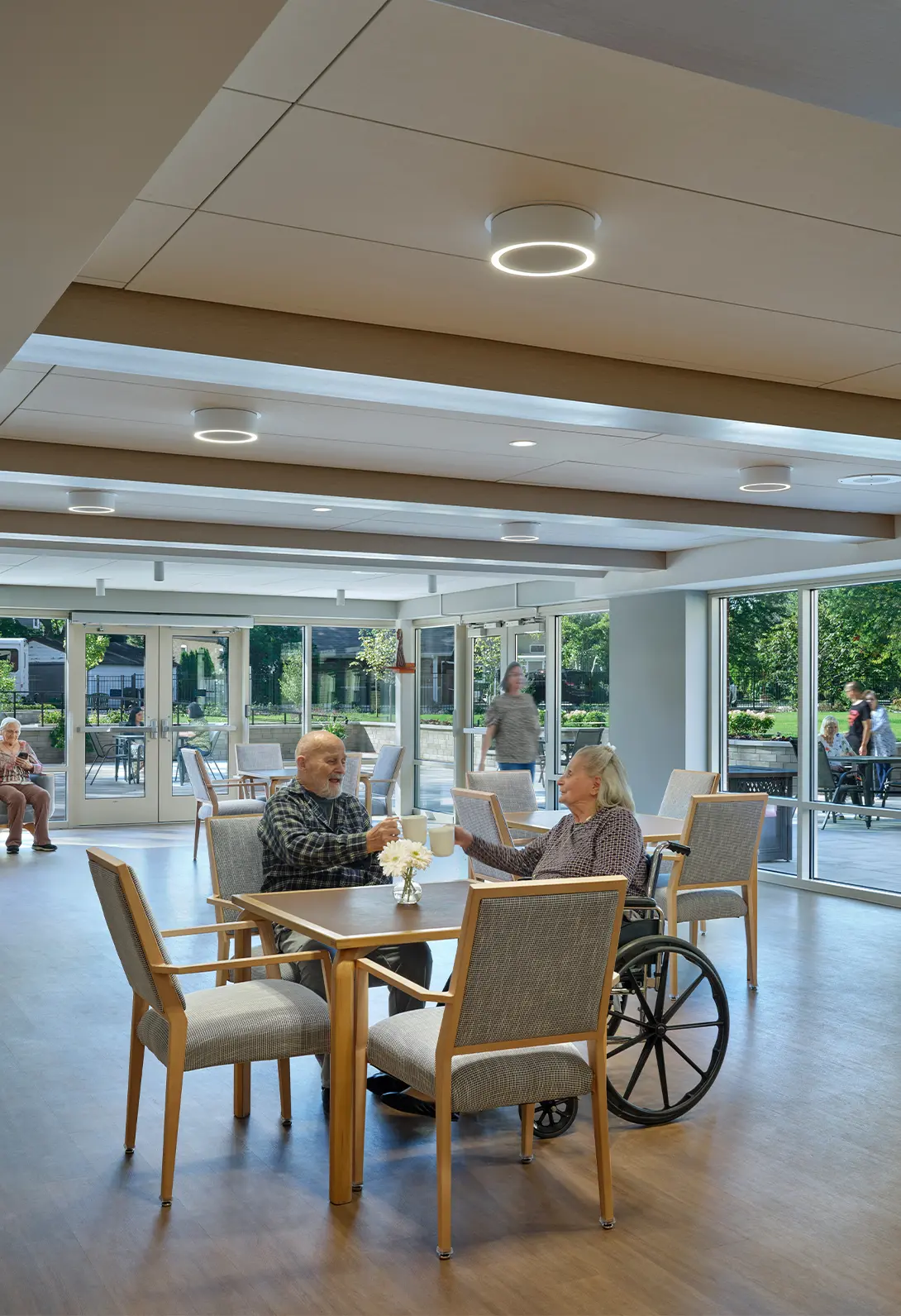 Elderly couple enjoying coffee in a bright, spacious social area with large windows and a garden view.