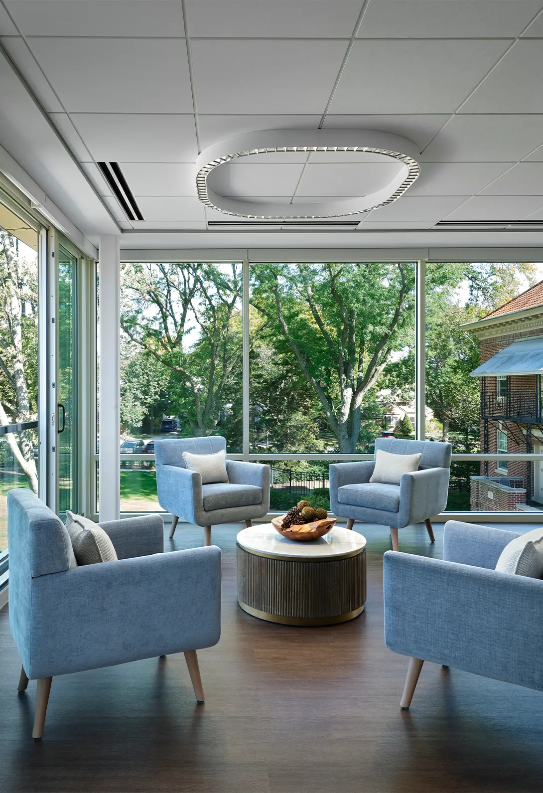 Modern lounge with large windows, four blue armchairs, and a round table with decor, overlooking a green landscape.