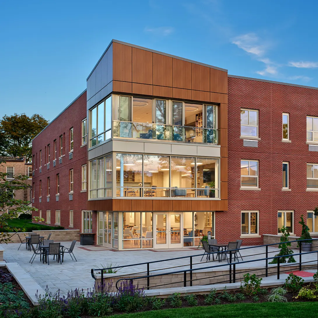 Modern brick building with large glass windows and a patio area, surrounded by greenery and a clear blue sky.