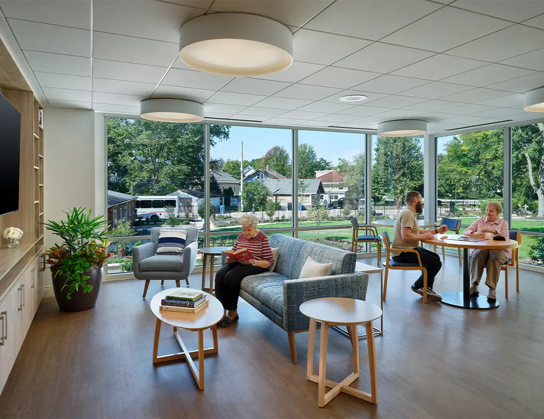 Modern senior living room with large windows, cozy seating, and people relaxing and chatting inside.