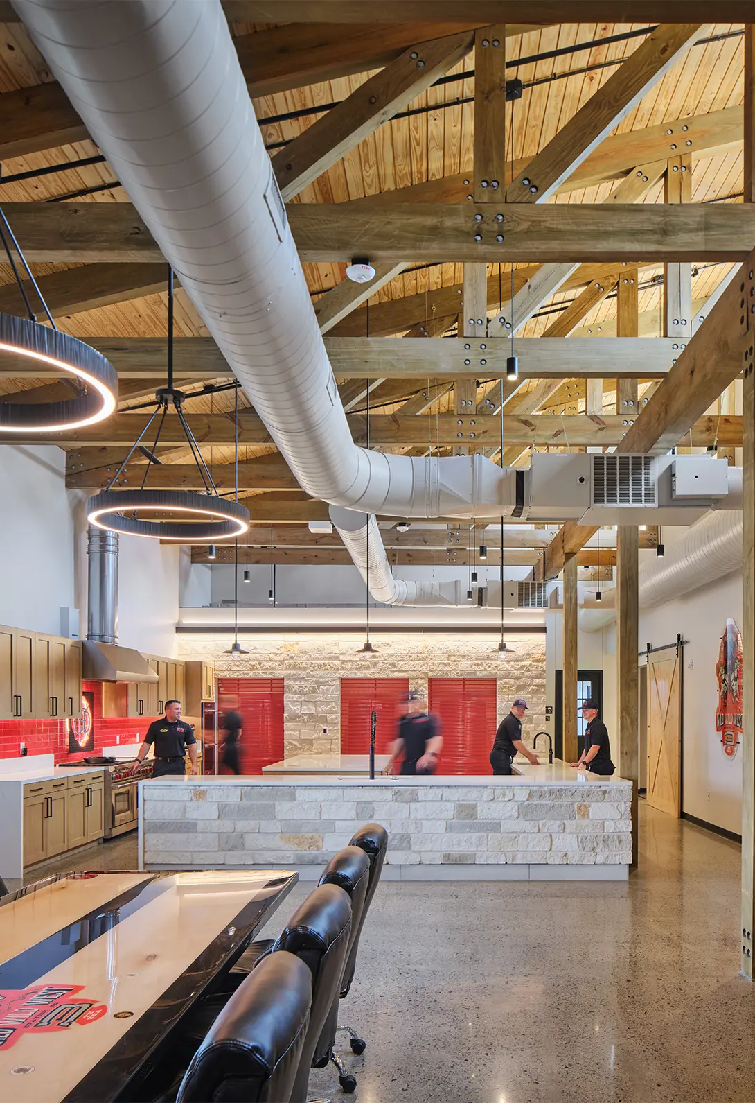Modern industrial kitchen with wooden beams, exposed ducts, and chefs preparing food at a stone island counter.