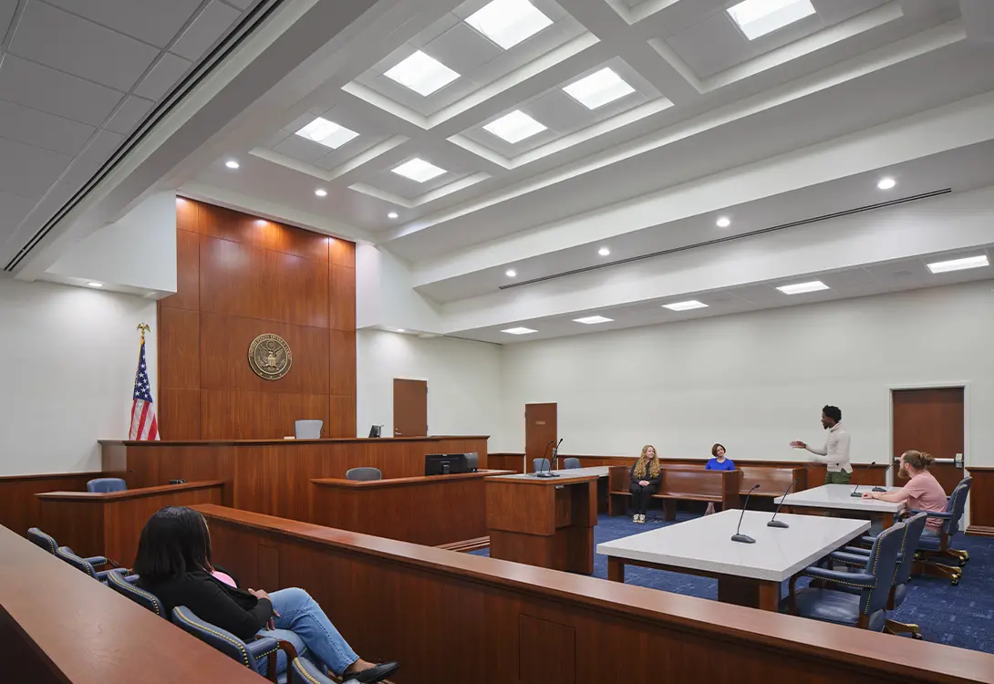 Modern courtroom scene with participants, empty jury box, and American flag under bright ceiling lights.