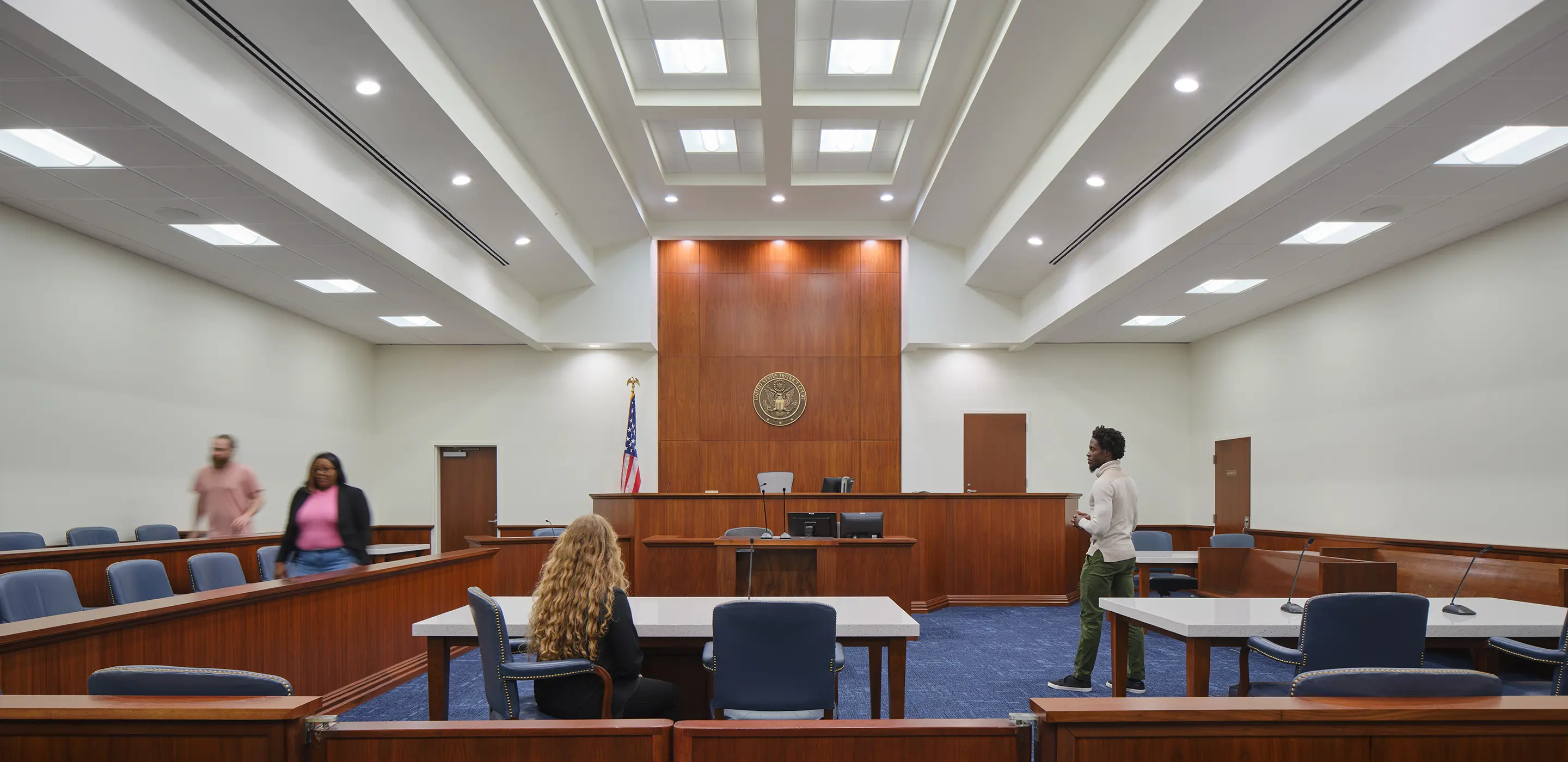 Modern courtroom interior with people present, showing wooden furniture, American flag, and bright ceiling lights.