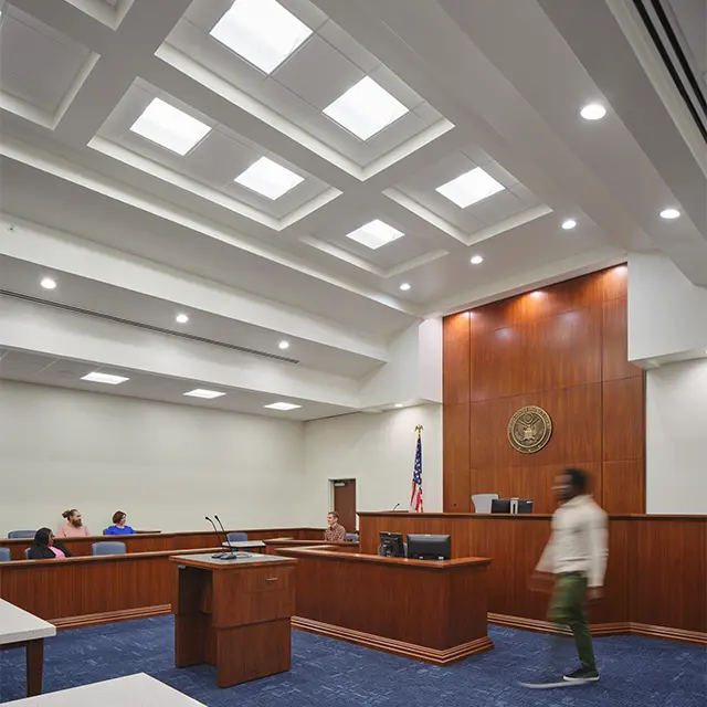 Modern courtroom interior with wooden paneling, people seated, and bright ceiling lights.