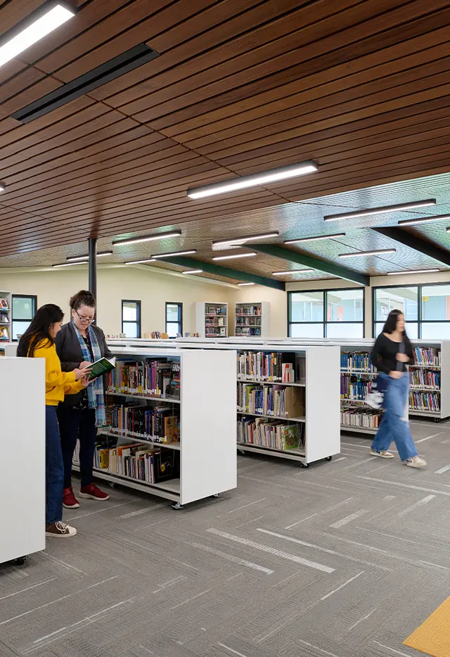 Modern library interior with people browsing bookshelves under wooden ceiling and bright lighting.