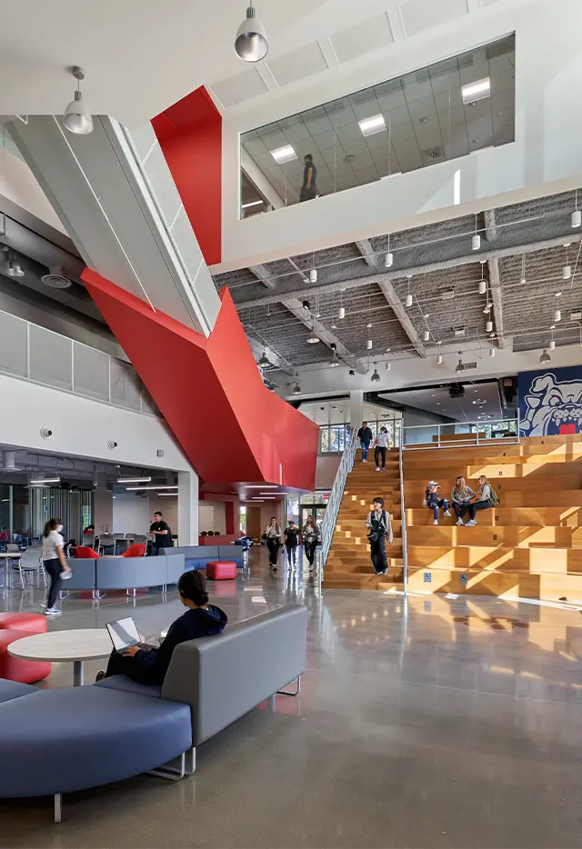 Modern university atrium with students on wooden steps, red architectural accents, and contemporary seating.