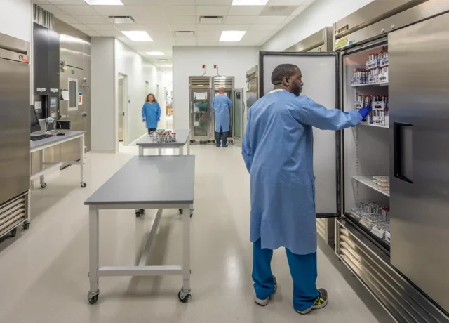 Laboratory staff in blue coats accessing samples from stainless steel refrigerators in a modern lab environment.