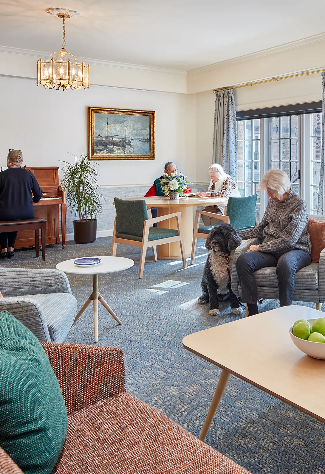 Cozy senior living room with people, a dog, and music, highlighting community and relaxation in a bright, furnished space.