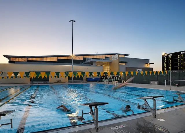 Outdoor swimming pool with divers and swimmers at sunset, with colorful flags and scoreboard visible.
