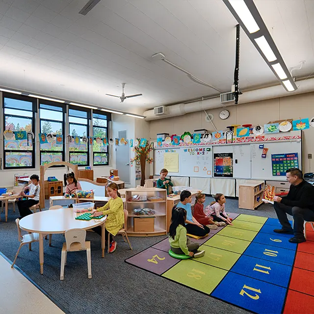 Children engaged in activities inside a colorful, well-lit classroom with a teacher reading to a group on the rug.