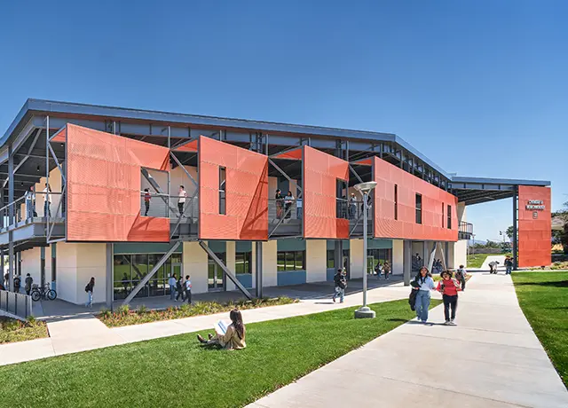 Modern educational building with orange exterior, located on a sunny campus, surrounded by students and green lawns.