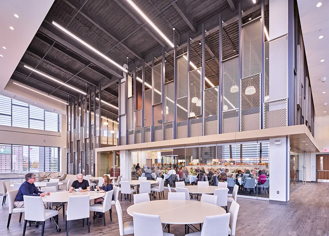 Modern cafeteria with people dining, featuring high ceilings, large windows, and contemporary interior design.