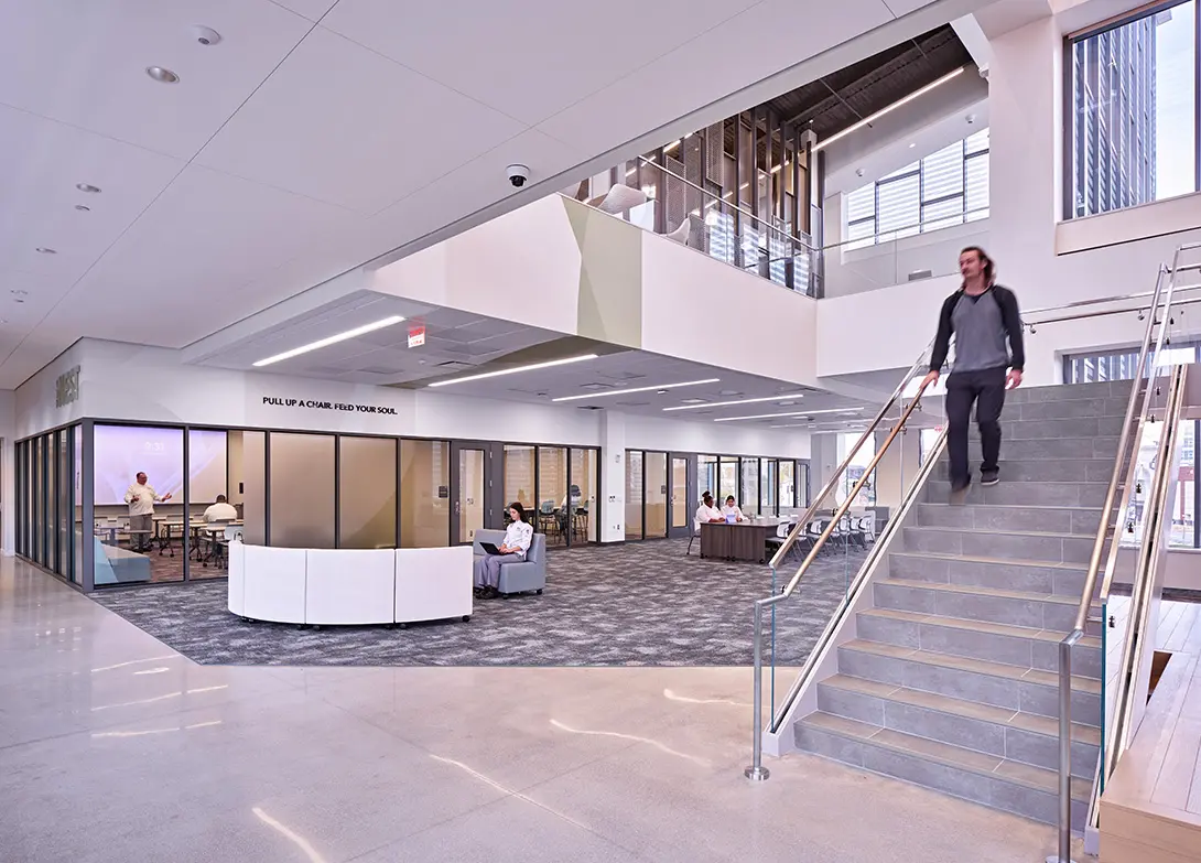 Modern office lobby with open seating, glass meeting rooms, and staircase. People working and walking in the space.