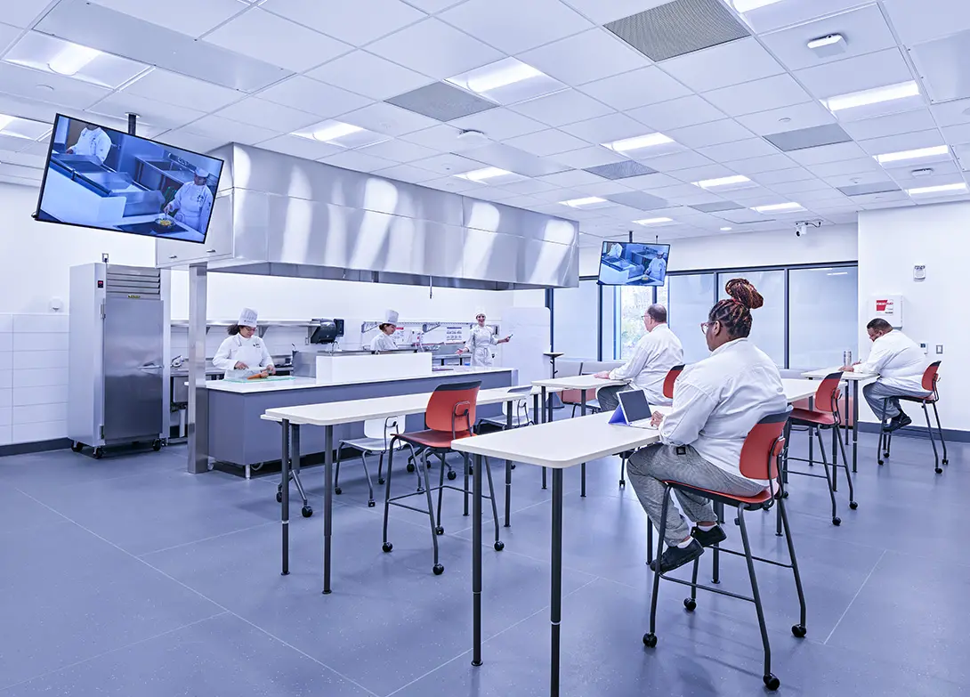 Modern culinary classroom with chefs in white uniforms and students observing cooking demonstrations on screens.