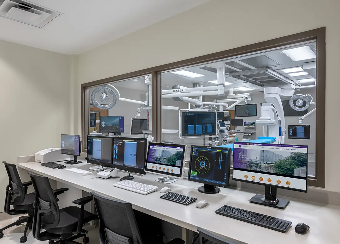 Modern medical control room with multiple monitors and office chairs, overlooking a high-tech operating room.