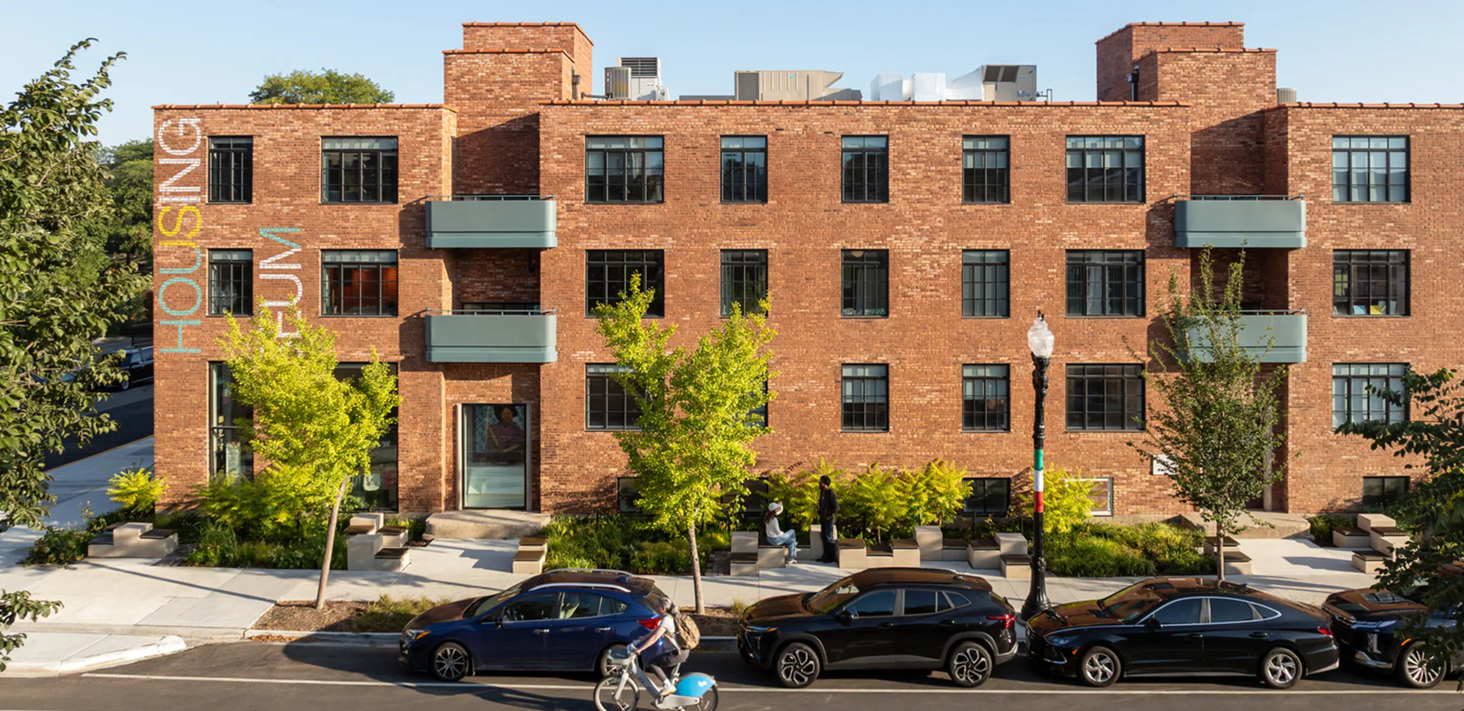 Red brick building with Housing sign, modern balconies, trees, and parked cars in a vibrant urban setting.