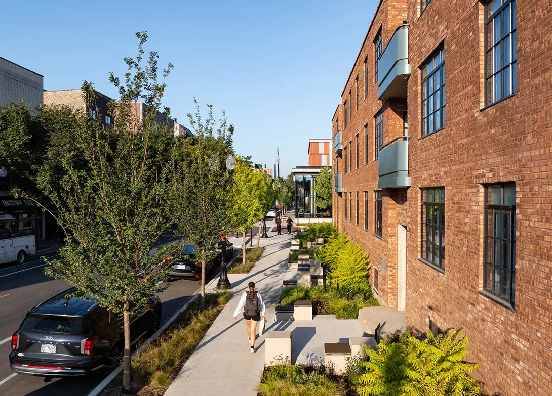 Modern urban street with a brick building, trees, and people walking on a sunny day.