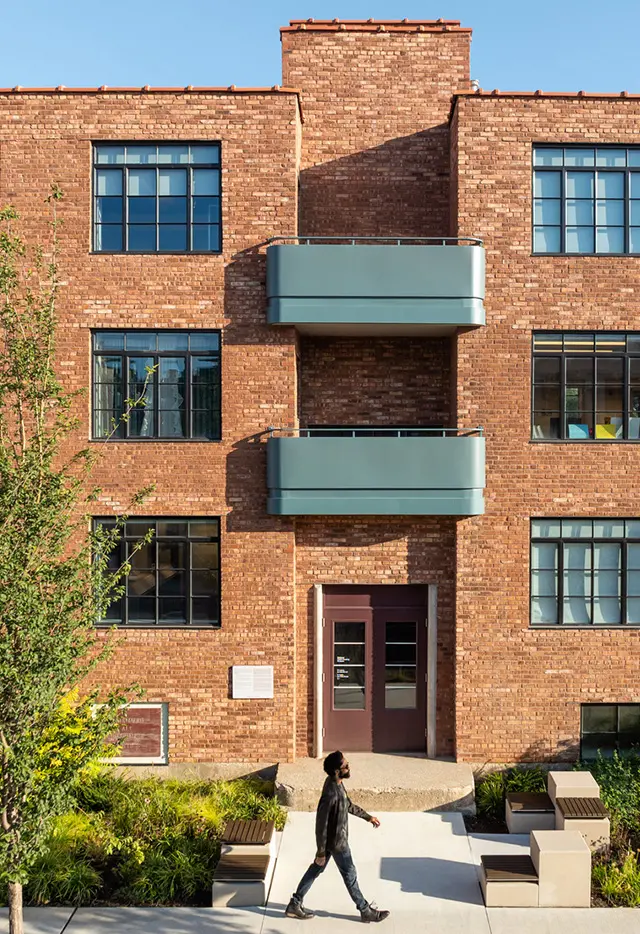 Person walking past a modern brick building with blue balconies and large windows on a sunny day.