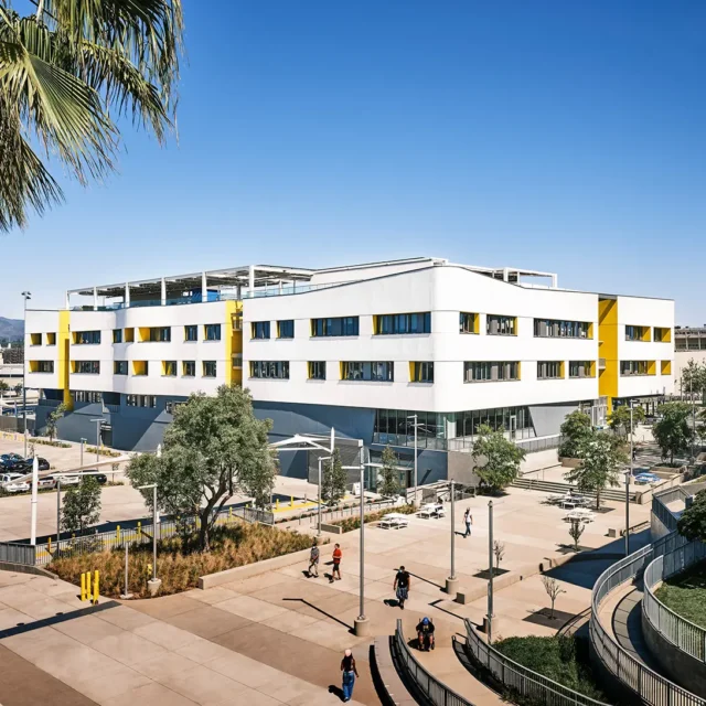 Modern educational building with yellow accents and outdoor walkway, surrounded by trees and people on a sunny day.