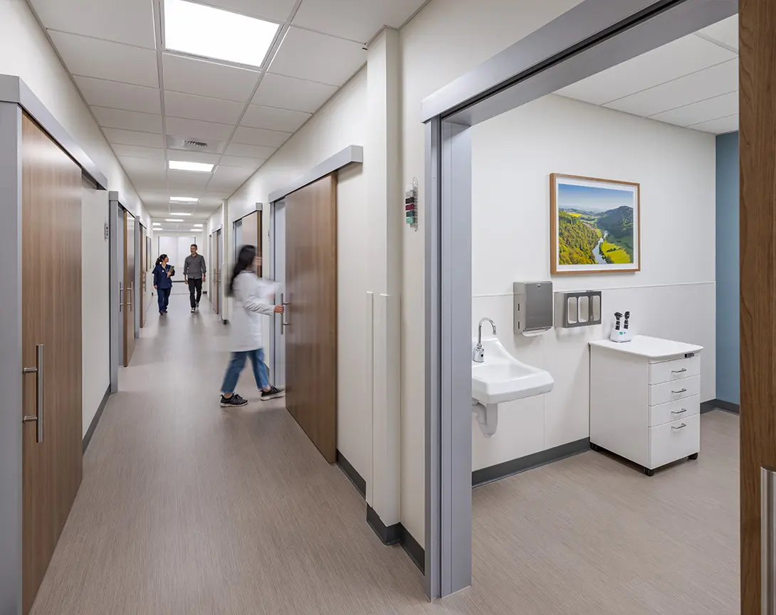 Modern hospital corridor with healthcare professionals, a sink area, and doors leading to patient examination rooms.