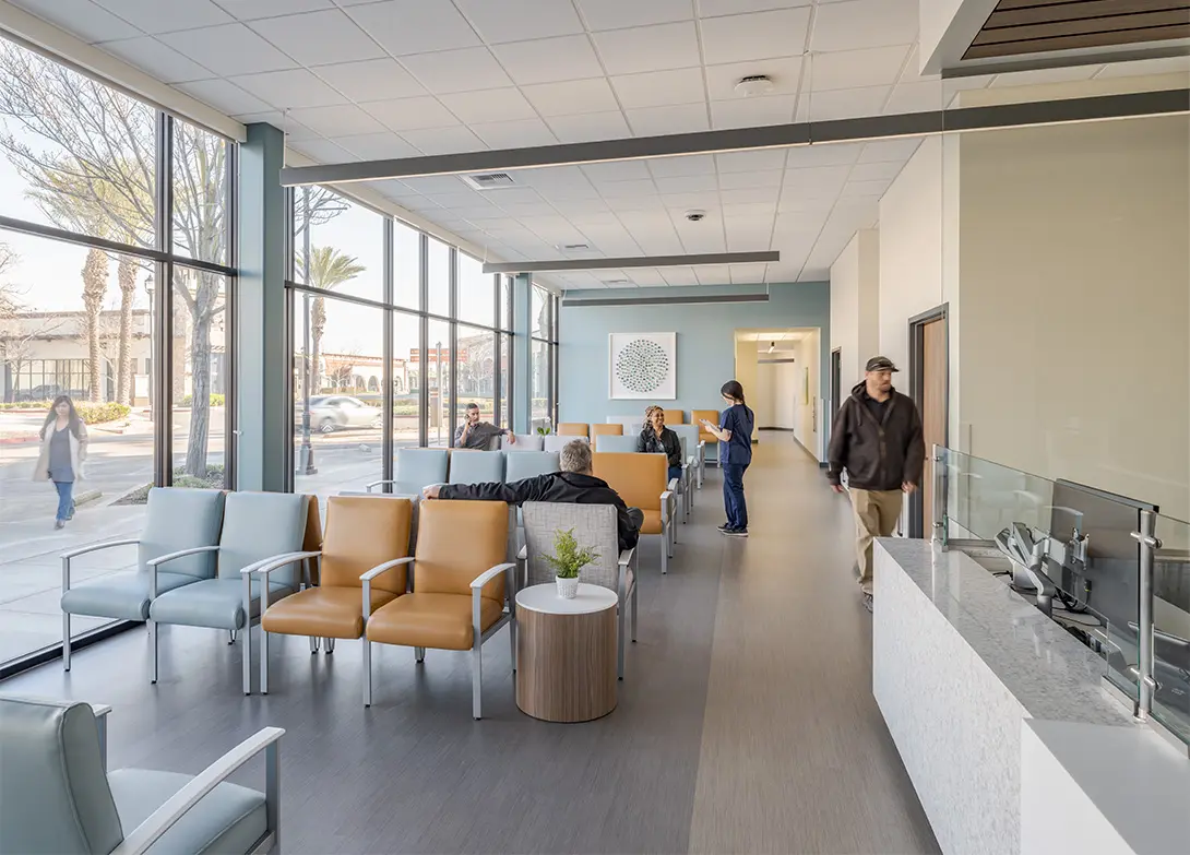 Modern clinic waiting area with large windows, comfortable chairs, and patients interacting with staff.