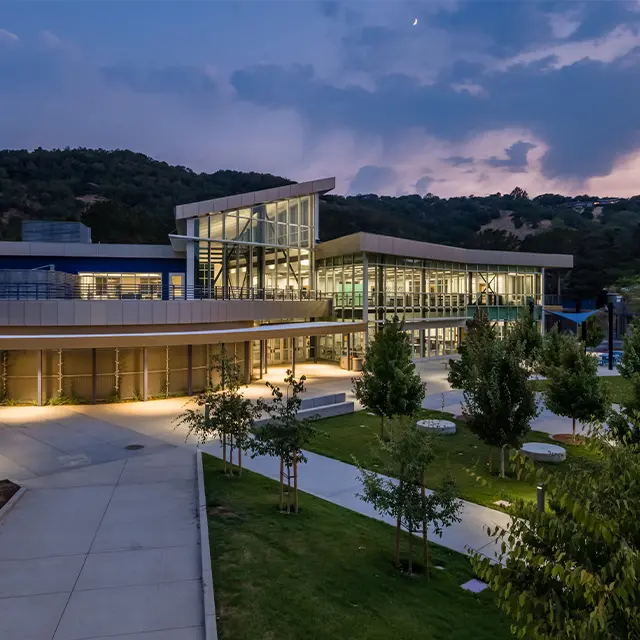 Modern building with glass facade and landscaped surroundings at dusk, set against a backdrop of hills and a colorful sky.