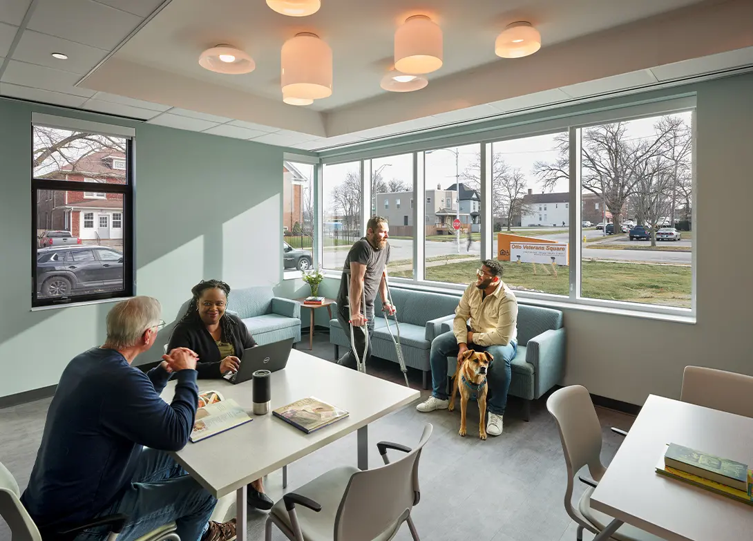 People in a bright community room chatting, with a dog by the window. Comfortable, inclusive space.