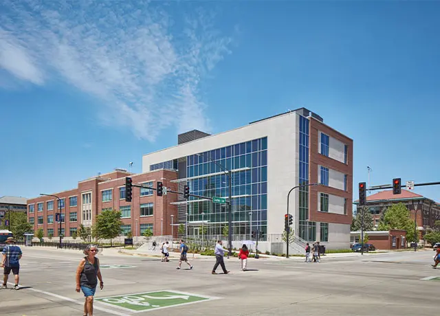 Modern urban intersection with pedestrians crossing near a large brick and glass building under a clear blue sky.