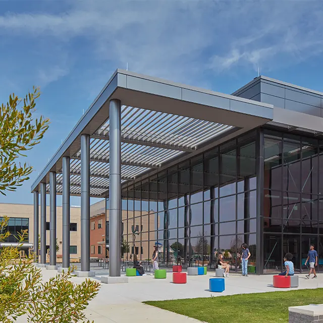 Modern glass-walled building with colorful outdoor seating under a spacious metal awning, people socializing.