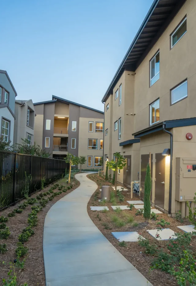 Modern apartment complex with a landscaped walkway, surrounded by greenery and contemporary architecture.