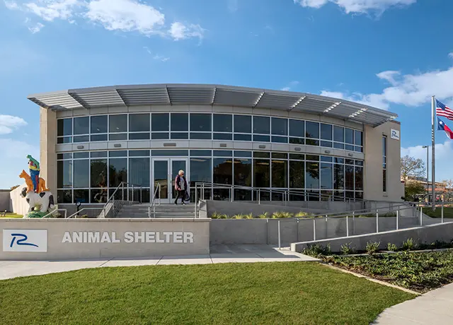 Modern animal shelter exterior with large windows, flags, and landscaped entrance, under a blue sky.