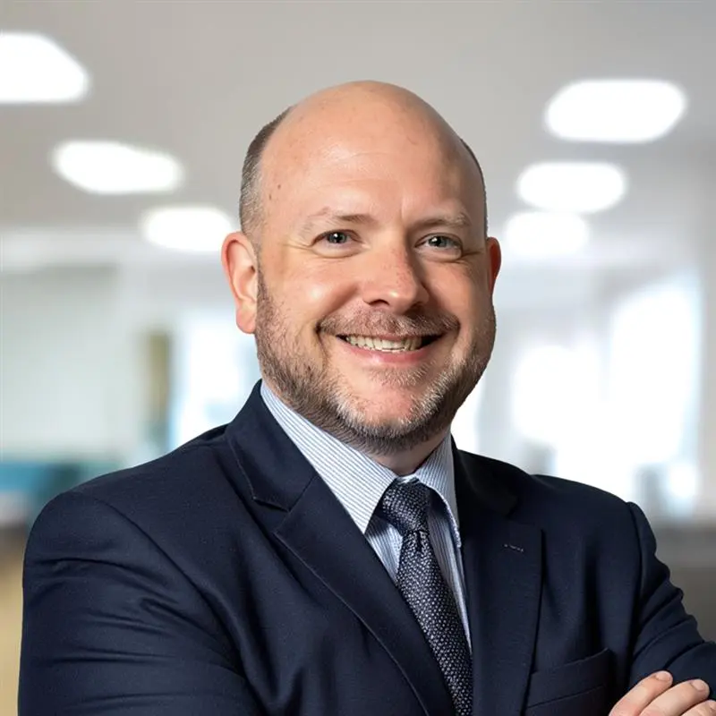 Smiling bald man in suit with arms crossed in a bright office setting.