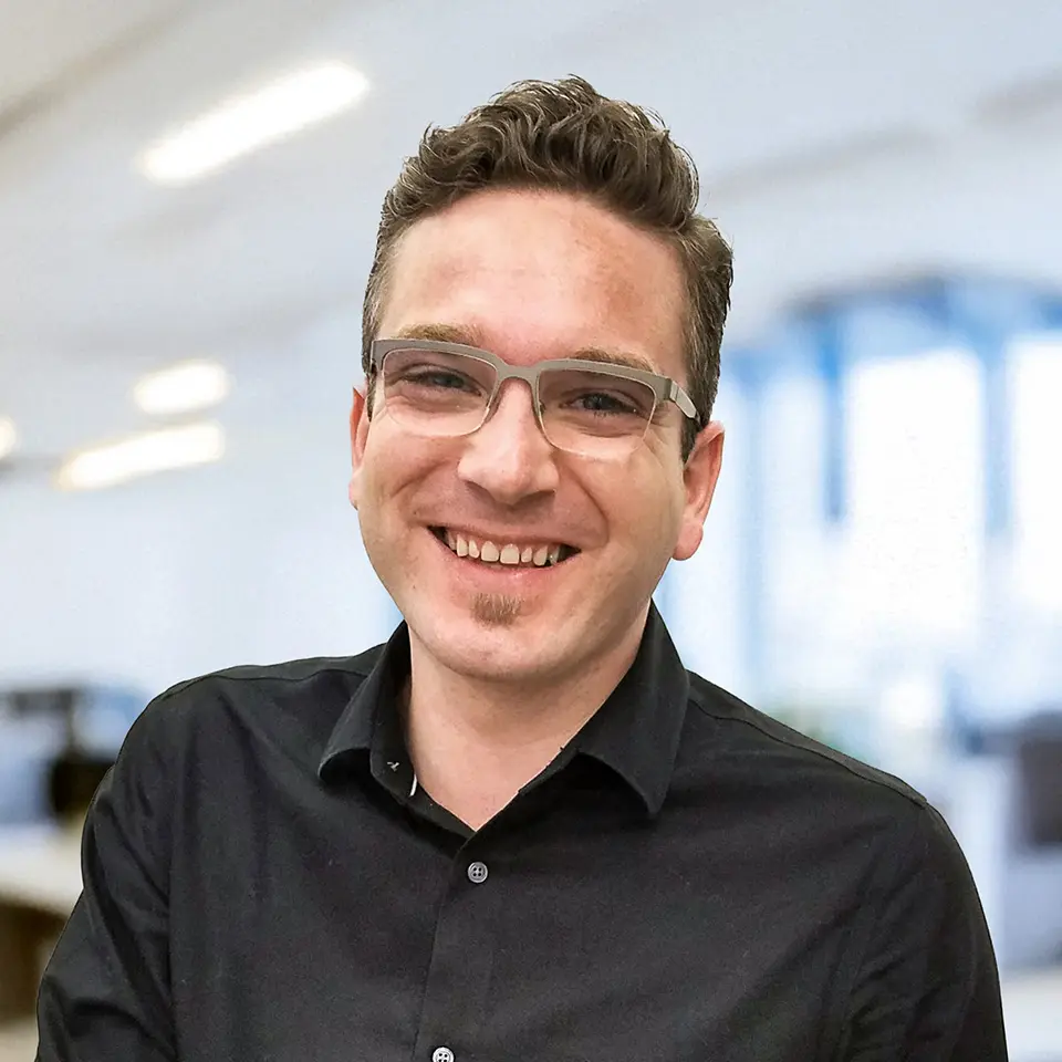 Smiling man in a black shirt with glasses, standing in a bright office setting.