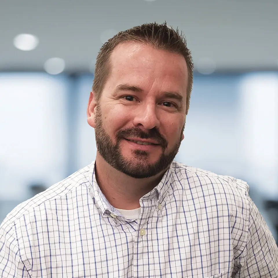 A smiling man in a checkered shirt with a blurred office background.