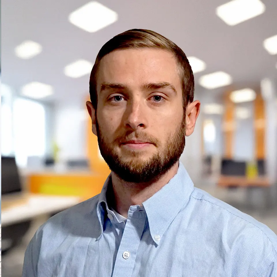 Man with beard in a blue shirt standing in a modern office setting with blurred background.