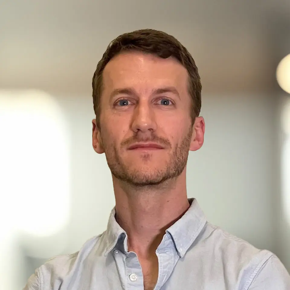 Portrait of a man in a light blue shirt with a neutral expression, soft-focus background.