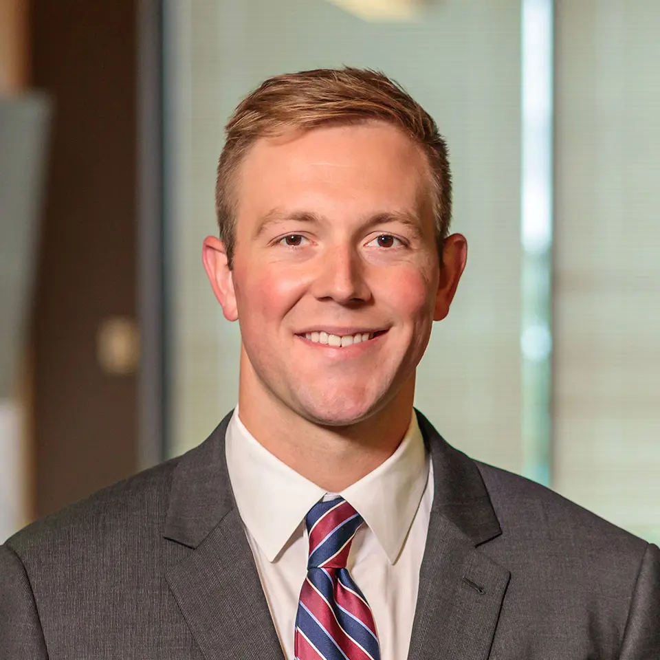 Portrait of a smiling man in a suit and tie, standing indoors with a blurred background.