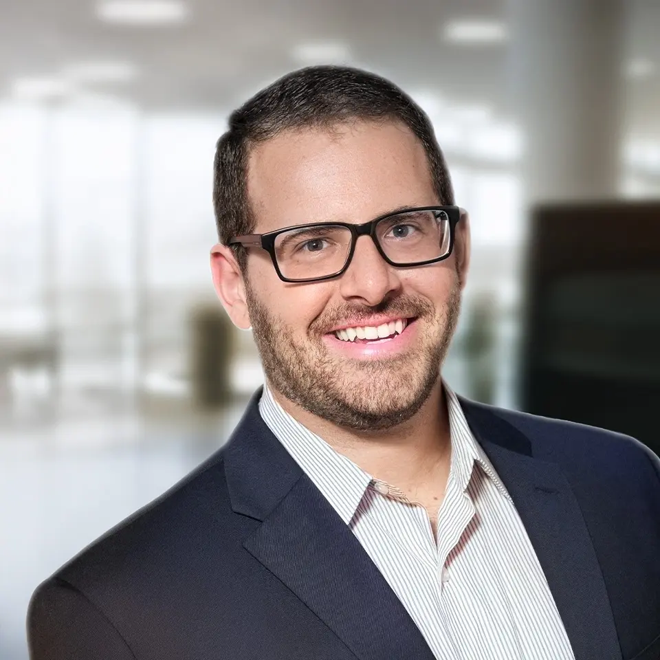 Smiling man with glasses and beard in office setting, wearing a suit jacket and striped shirt.
