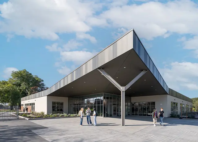 Modern building with geometric roof design under a blue sky, surrounded by greenery and a few people walking.